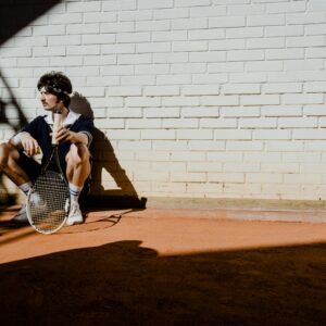 A stylish man in vintage sportswear sits with a tennis racket and balls, casting shadows.